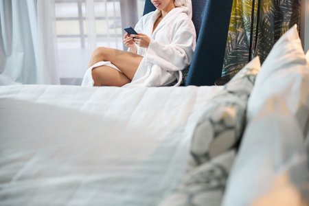 Cropped copy-space photo of a mirthful young woman in white bathrobe looking at cellphone screen while sitting in arm-chairの写真素材