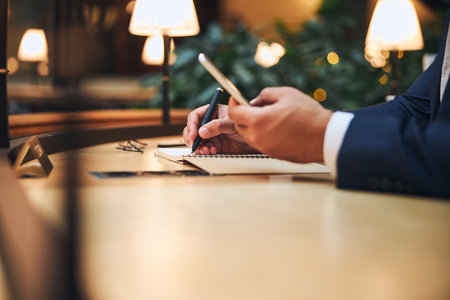 Cropped photo of a businessman with a cellphone in the hand taking notes in his notebook at the...