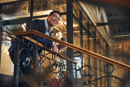 Smiling bespectacled man with a smartphone and a beverage in his hands gazing into the distanceの写真素材