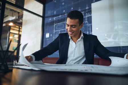 Waist-up portrait of a smiling pleased male employee studying a technical drawing at the deskの写真素材