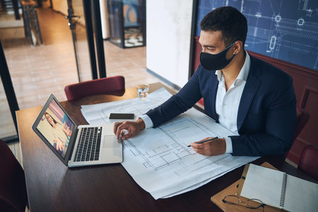 Young male engineer in a face mask communicating with his coworker during the video callの写真素材