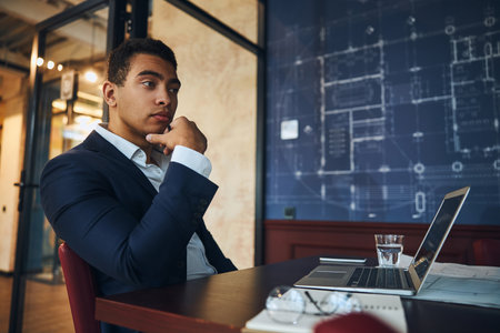 Waist-up portrait of a good-looking thoughtful male office worker pondering in front of his laptopの写真素材