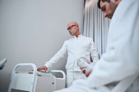 Low angle of bald doctor in medical glasses using equipment for treating young man in clinic officeの写真素材