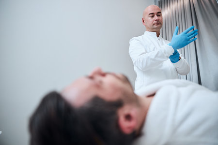 Low angle of bald male doctor wearing rubber gloves while working in clinic with handsome patientの写真素材
