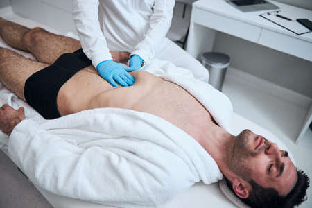 Top view of stubbled handsome guy lying on couch while doctor is touching his abdomen with handsの写真素材