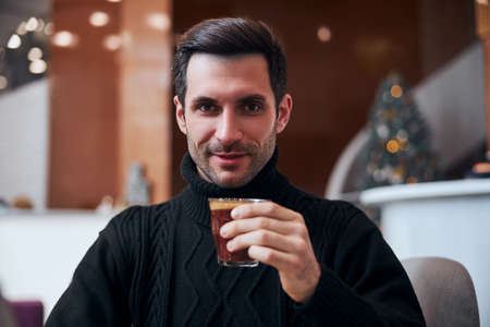 ortrait of smiling handsome male sitting in lobby during Christmas holidays and drinking cup of...