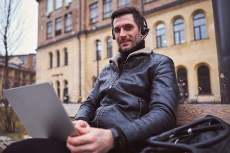 Low angle waist up portrait of smiling stubbled guy sitting on bench and using laptop and earphones