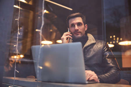 Young guy is sitting at window in bar and talking on cell phone while surfing internet on notebook