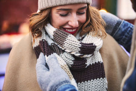 Smiling female is having fun while her caring partner is putting on warm scarf in city centreの写真素材
