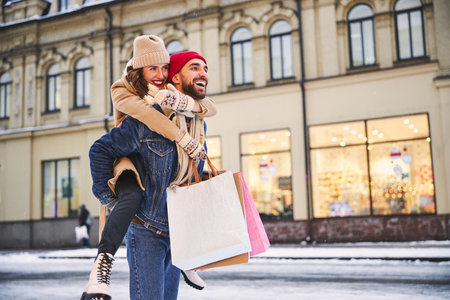 Happy man is carrying girlfriend and paper bags while buying presents in city for winter holidaysの写真素材