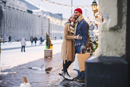 Cheerful bearded guy is embracing pretty girlfriend while going shopping together outdoors during Christmas holidaysの写真素材