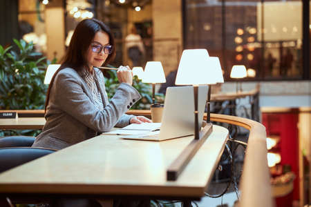 Cute woman wearing glasses while looking at screen of laptop and making notes