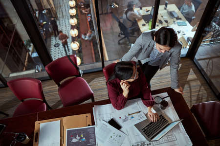 Serious Asian female sitting at her workplace while listing to her colleague