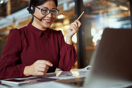 Kind international female person expressing positivity while using her headphones for online workの写真素材