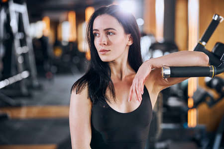 Cheerful pretty athletic woman is relaxing at sport machine during break in strength workouts with equipmentの写真素材