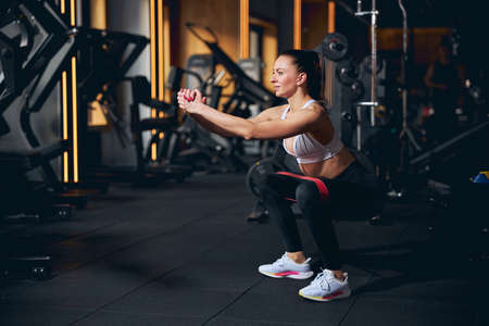 Jolly pretty slim female is exercising lower body with resistance band in modern sport club