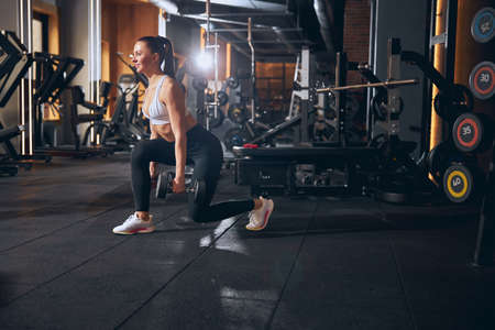 Smiling fit pretty female is training legs and lifting heavy dumbbells during visit to gym