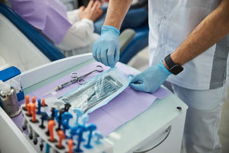 Man carefully opening a poly bag, lying on a medical table near a rack with dental composite resinsの写真素材