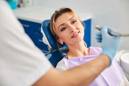 Middle-aged lady resting on dental chair while looking at handpiece in doctor hand moving to her teethの写真素材