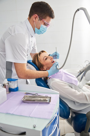 Dentist in a facial mask treating the teeth of a female under operation by using dental toolsの写真素材
