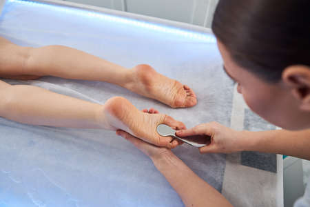 Massage practitioner pressing a stainless steel scraper tool to the sole of female left foot during a beauty procedureの写真素材