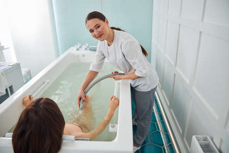 Female worker of spa facility pointing a jet of water from an elastic tube to waist of a female