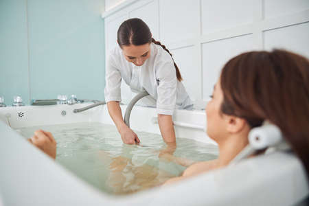 Woman in white shirt with brown ponytail leaning over an edge of bathtub for placing tube close to...
