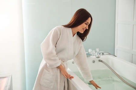 Attractive lady in white bathrobe looking at water and smiling while planning to take revitalizing bath in spa salonの写真素材