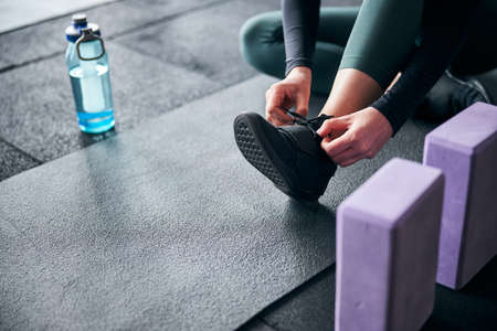Croppe photo of a lady tying her shoelaces on a black sneaker whie sitting on sports matの写真素材