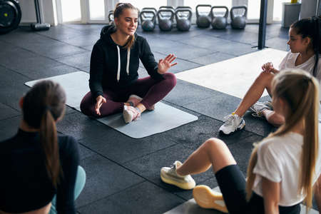 Young fitness specialist sitting on gym floor with young women and having a seminar on working outの写真素材