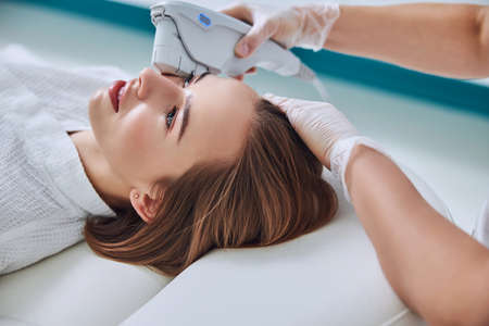 Charming young woman lying on medicine chair while unrecognized beautician doing spa treatment in spa centerの写真素材