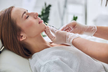 Charming Caucasian young woman with close eyes lying on medicine chair while getting hyaluronic acid skincare in beauty clinicの写真素材