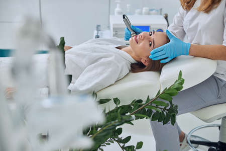 Charming funny Caucasian female lying on medicine chair while receiving rejuvenation facial procedure in beauty clinicの写真素材