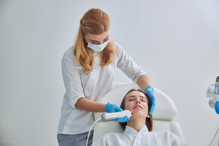 Young adorable redheaded woman in white bathrobe receiving professional anti aging skin procedure in beauty clinicの写真素材
