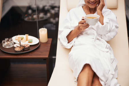 Charming beautiful elegant woman in white bathrobe drinking beverage while spending time in spa...
