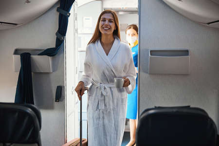 Relaxed female coming inside plane cabin in robe with coffeeの写真素材