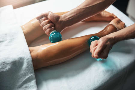 Beautiful elegant feet lying on massage table while male masseur doing massage with cold glass in health clinicの写真素材