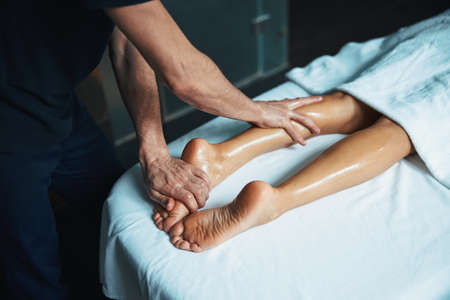 Charming young woman legs on white towel and man hands healing body treatment in beauty clinicの写真素材