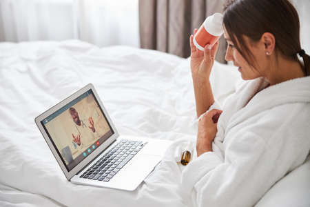 Caucasian woman holding orange pill jar in hand while looking to the screen of computerの写真素材