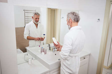 Handsome bearded male looking to the bottle with cosmetic in front of the mirror in bathroomの写真素材