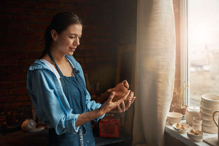 Beautiful young woman ceramic sculptor looking to the lump of earthenware product in arms in pottery...