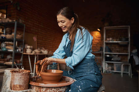 Young Caucasian charming ceramicist wearing apron modeling clay bowl in pottery workshopの写真素材