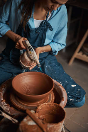 Focused Caucasian female artisan sitting near the pottery wheel while making tableware in pottery workshopの写真素材