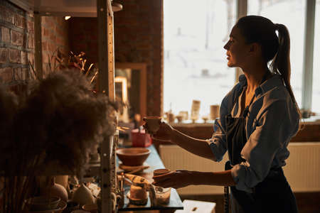 Beautiful Caucasian woman with clay tableware in hands standing near the shelf in art studio