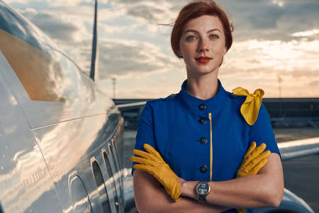 Calm red-haired stewardess posing for the camera by an aircraft