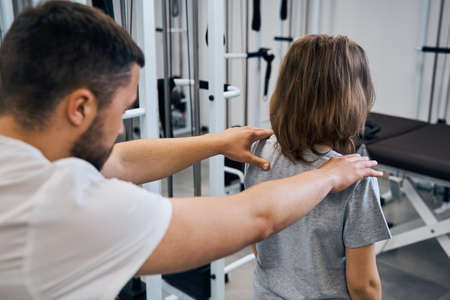 Massage therapist treats girl in medical rehab center back view. Chiropractor checks spine, neck