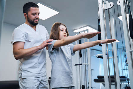 Pediatric therapist checks posture of girl with her arms stretched forward in rehabilitation center