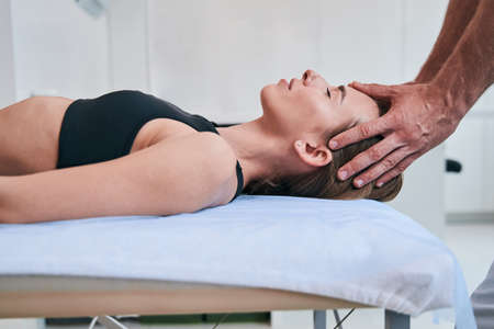 Young elegant Caucasian woman in black sports bra enjoying facial massage in medical office