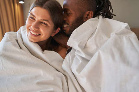 Romantic mixed-race couple wrapped in duvets sitting in bedの写真素材