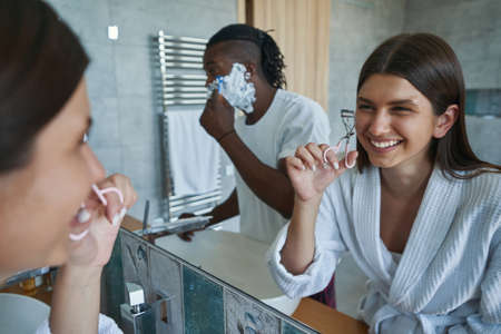 Joyous lady curling her eyelashes while guy shaving his faceの写真素材